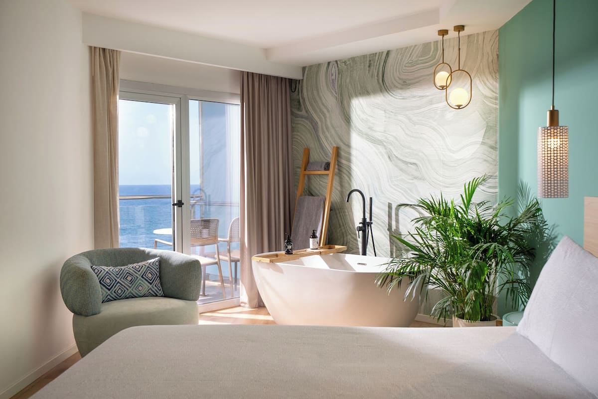 A modern hotel room with a large bed, a bathtub, a balcony with a view, and a potted plant.