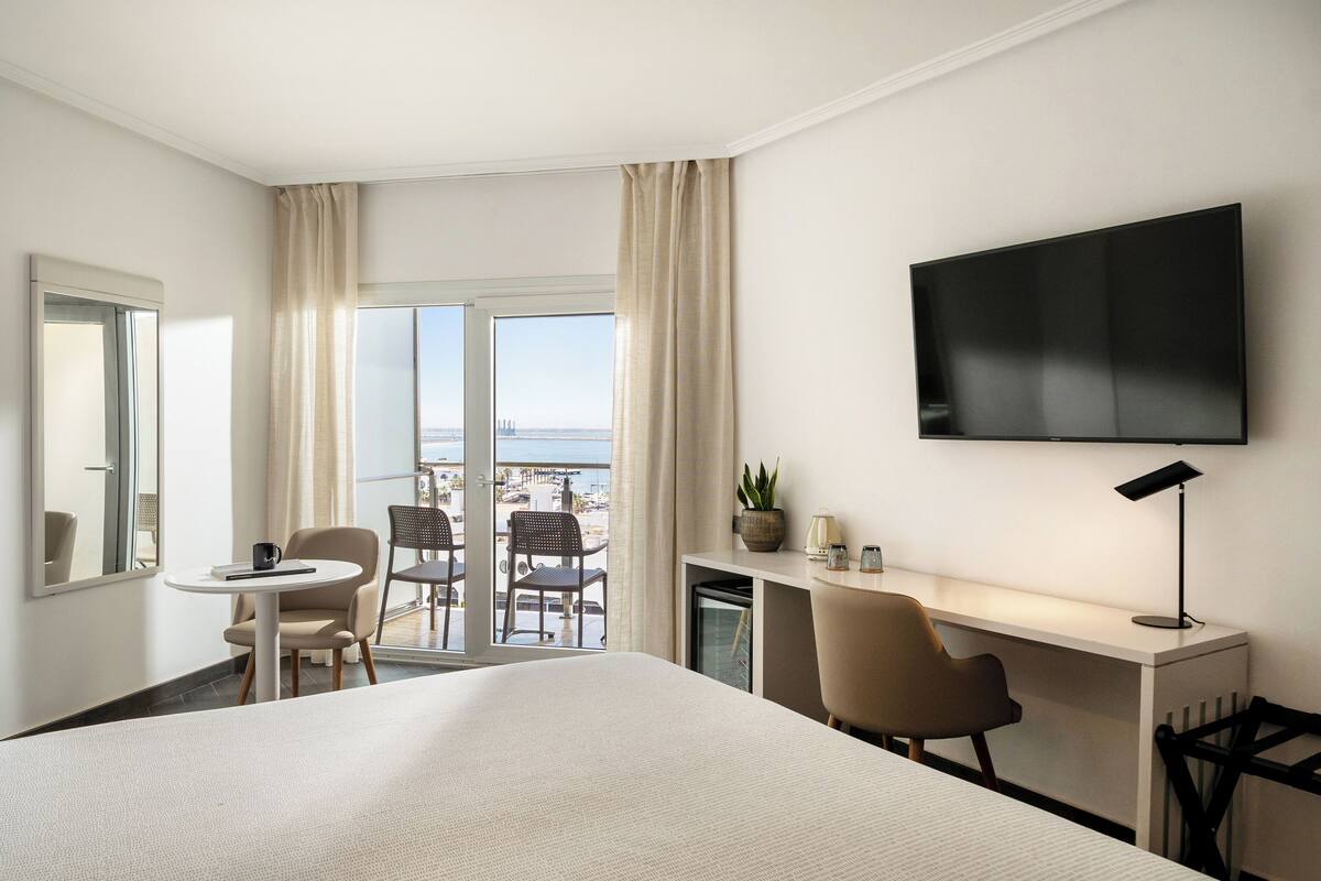 A hotel room with a large bed, a desk with a TV, a small table, two chairs, and a view of the city through the window.