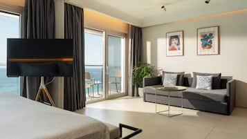 Habitación de hotel moderna con una cama grande, un sofá, una mesa de centro y una televisión. Incluye un balcón con vista al mar.