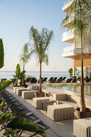 A poolside area with lounge chairs, striped ottomans, and potted plants.