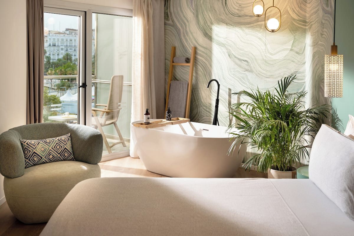 A modern bedroom with a large bed, a sitting area with a chair, a bathtub, and a potted plant.