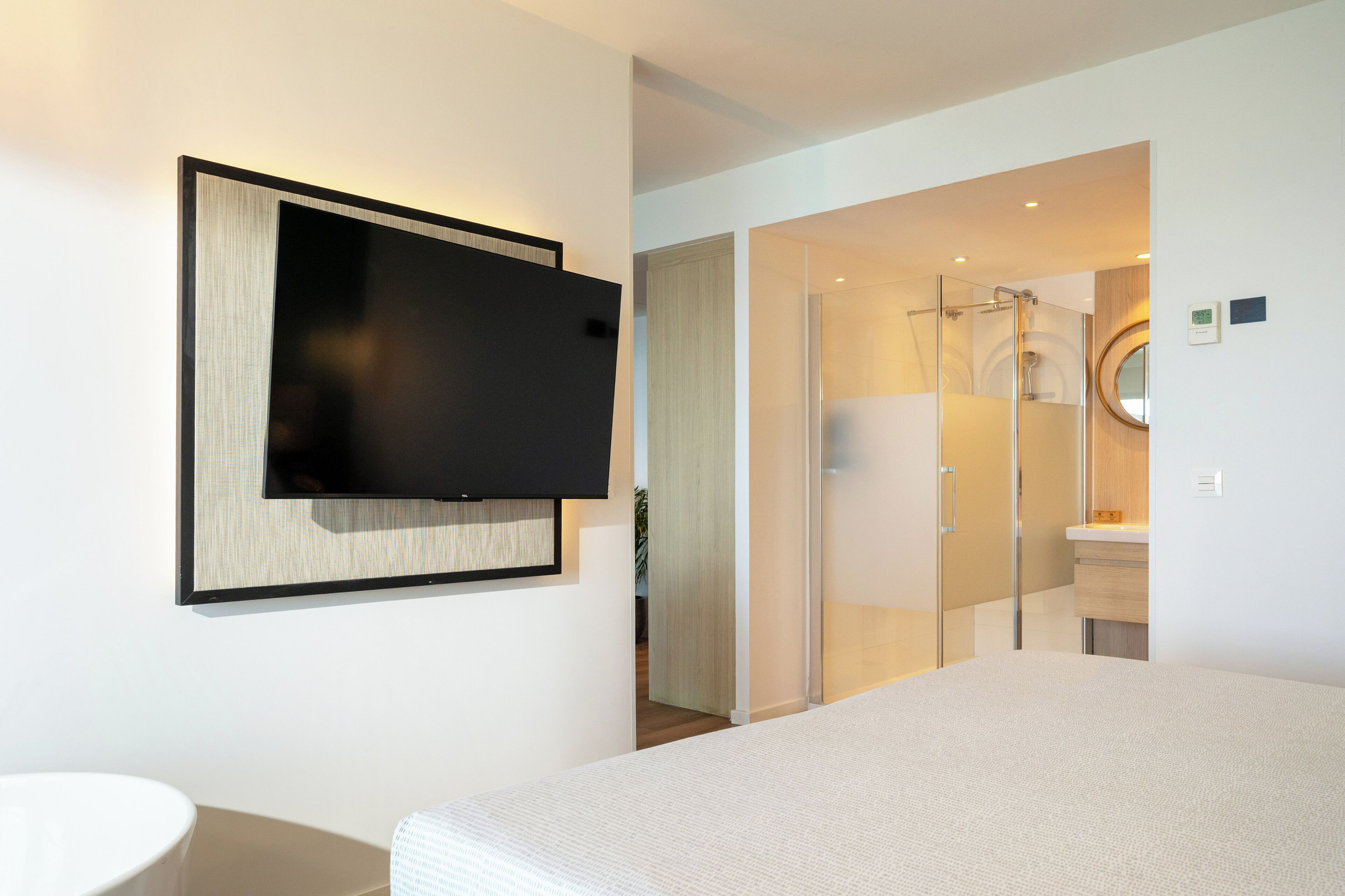 Quarto de hotel moderno com uma cama grande, TV de tela plana na parede, box de chuveiro de vidro e banheiro pequeno com espelho e pia.