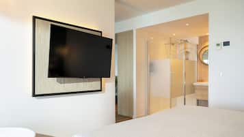 Quarto de hotel moderno com uma cama grande, TV de tela plana na parede, box de chuveiro de vidro e banheiro pequeno com espelho e pia.