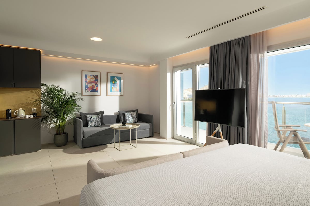 A modern hotel room with a large bed, a sofa, a TV, and a balcony with a view of the sea.