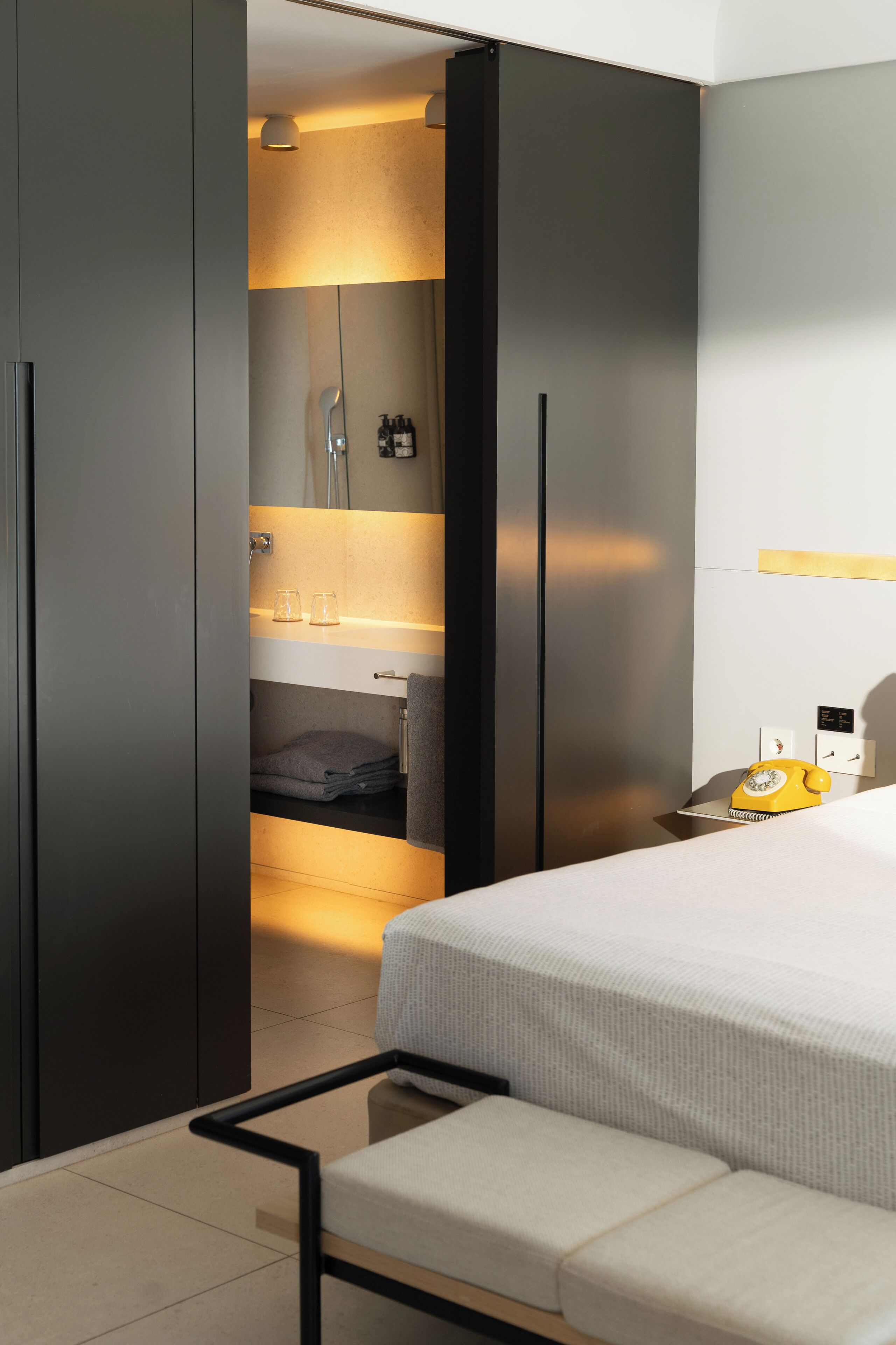 Ein modernes Hotelzimmer mit Bett, einer Sitzbank, einem Kleiderschrank, einem Spiegel, einem Waschbecken und Pflegeprodukten.