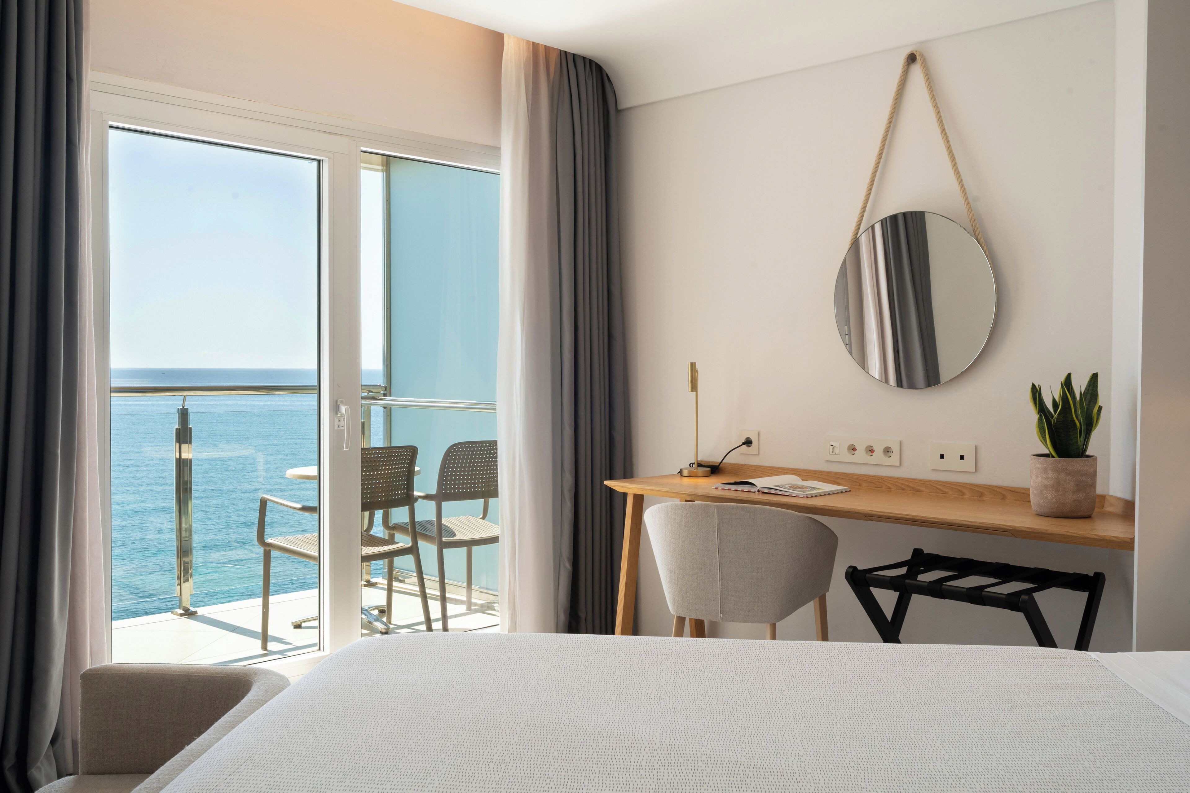 Habitación de hotel con cama, escritorio con silla, planta en maceta, espejo redondo y vista al mar a través de una puerta corredera.