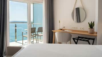 A hotel room with a bed, a desk with a chair, a potted plant, a round mirror, and a view of the sea through a sliding door.