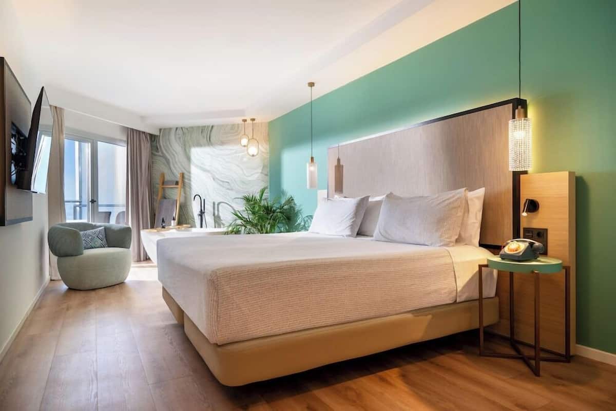 A modern bedroom with a large bed, a side table, a chair, and a view of the outdoors.
