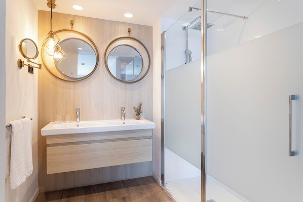 Un baño moderno con tocador de madera, dos espejos redondos, cabina de ducha de vidrio y un perchero para toallas.