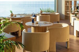 Un comedor moderno con mesas y sillas redondas, vista al océano y una estantería de fondo.
