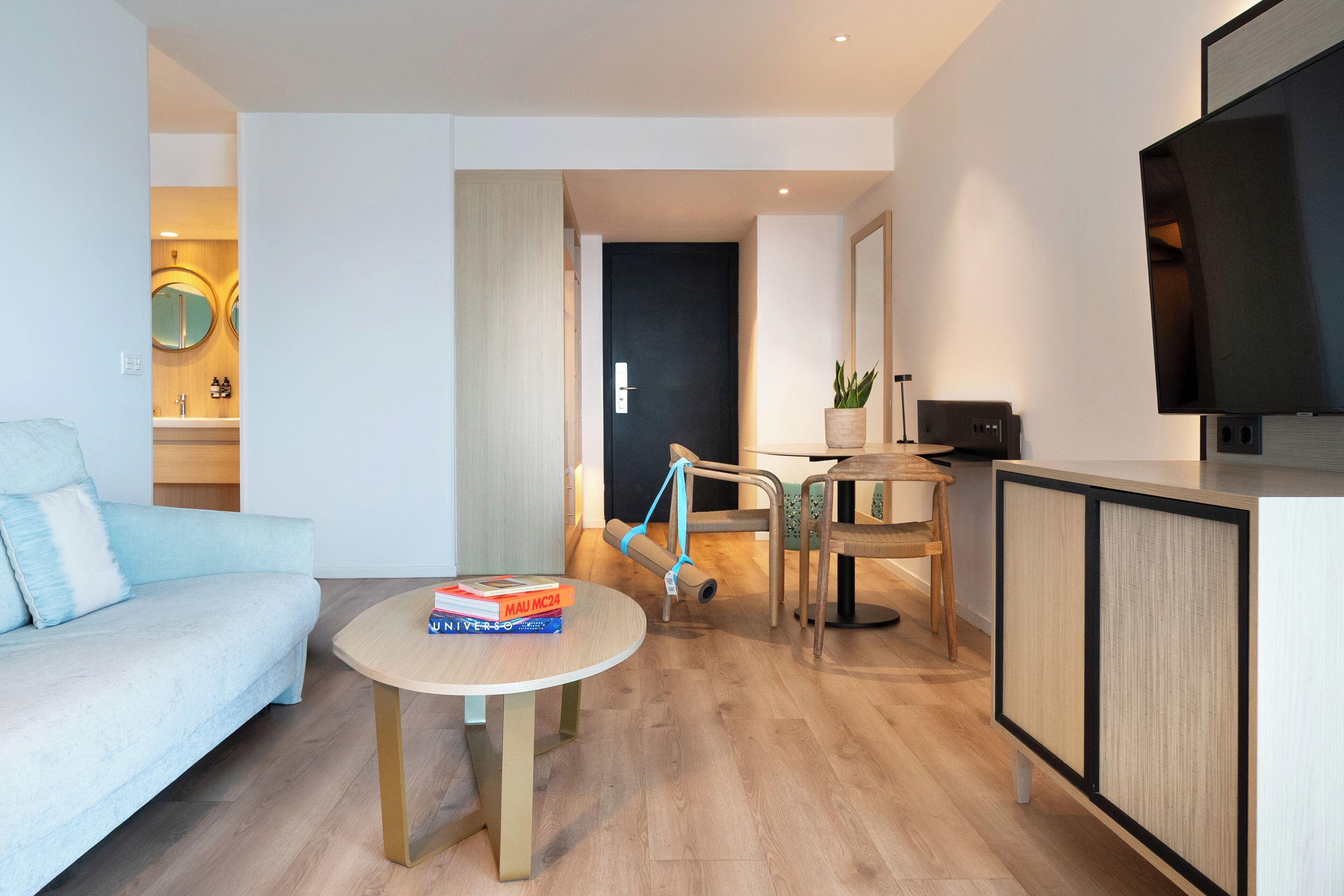 Una sala de estar moderna con un sofá azul claro, una mesa redonda con libros, un televisor sobre un mueble bajo y piso de madera.