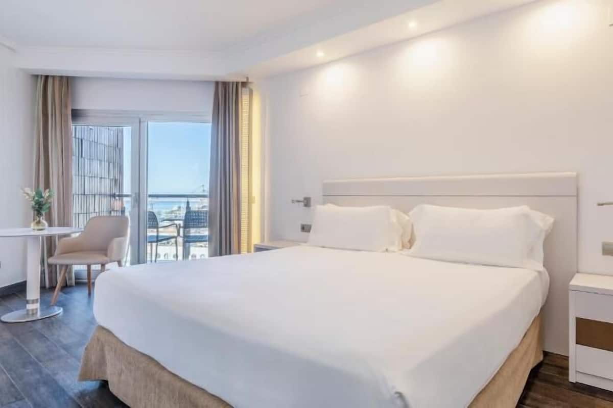 A hotel room with a large bed, a small table, a chair, a nightstand, and a view of the outside.