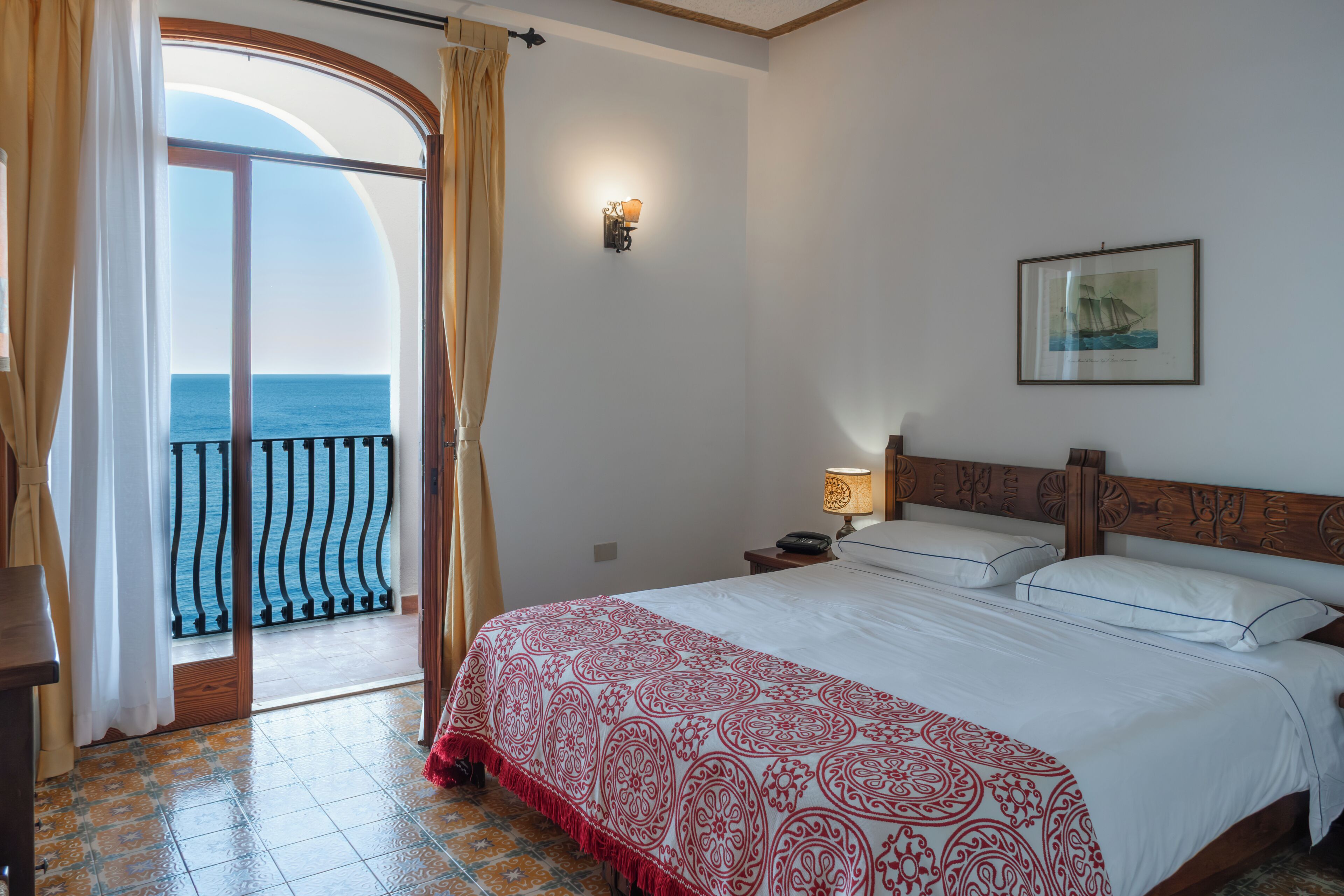 Une chambre d’hôtel avec un lit, une table de chevet, une lampe, un tableau encadré au mur et une vue sur la mer par une porte vitrée.
