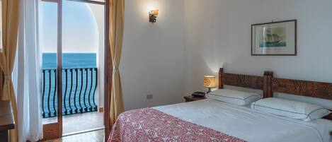 Une chambre d’hôtel avec un lit, une table de chevet, une lampe, un tableau encadré au mur et une vue sur la mer par une porte vitrée.