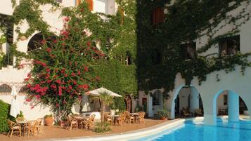 Zona de piscina con una piscina azul cristalina, rodeada de edificios blancos con balcones y contraventanas rojas.