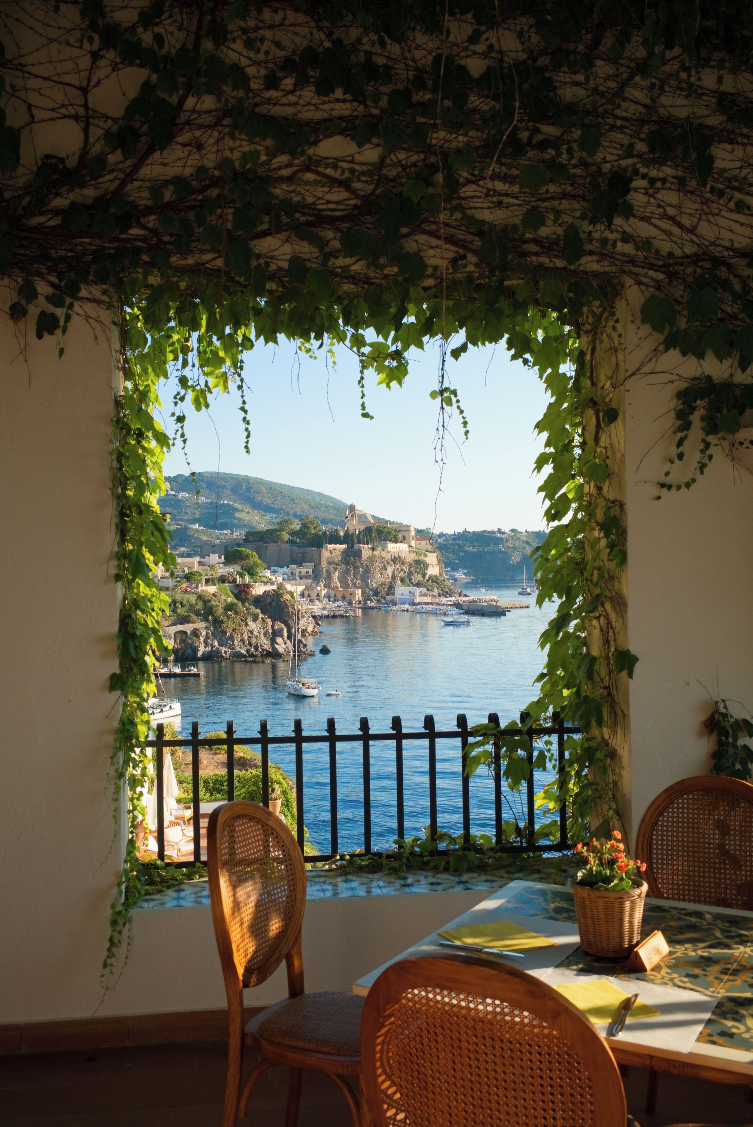 A dining area with a view of a coastal town, featuring a table with chairs, a view through a vine-covered window, and a railing.