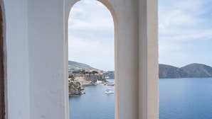 A coastal view through an arched window, featuring a harbor with boats, a small town, and hills in the background. - Hotel Carasco (Lipari)