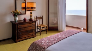 A hotel room with a bed, a dresser, a chair, a mirror, a lamp, and a view of the sea through a large window. - Hotel Carasco (Lipari)