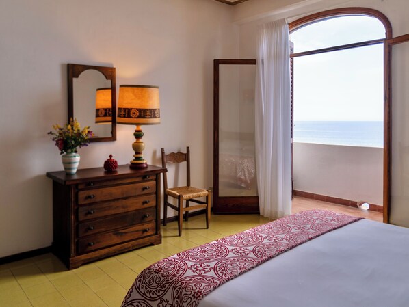A hotel room with a bed, a dresser, a chair, a mirror, a lamp, and a view of the sea through a large window. - Hotel Carasco (Lipari)