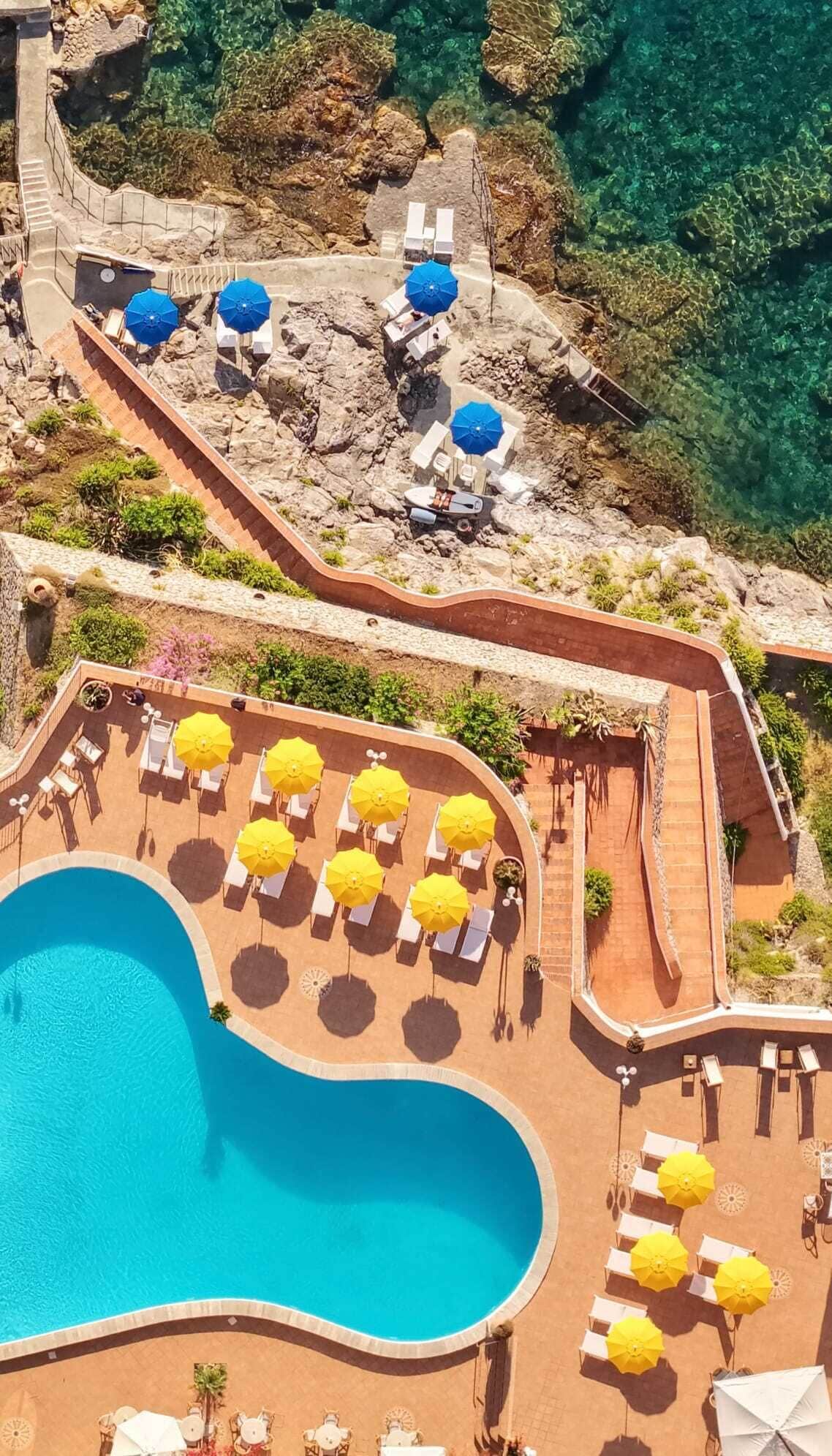 A pool area with lounge chairs and umbrellas, surrounded by a rocky landscape and clear water.