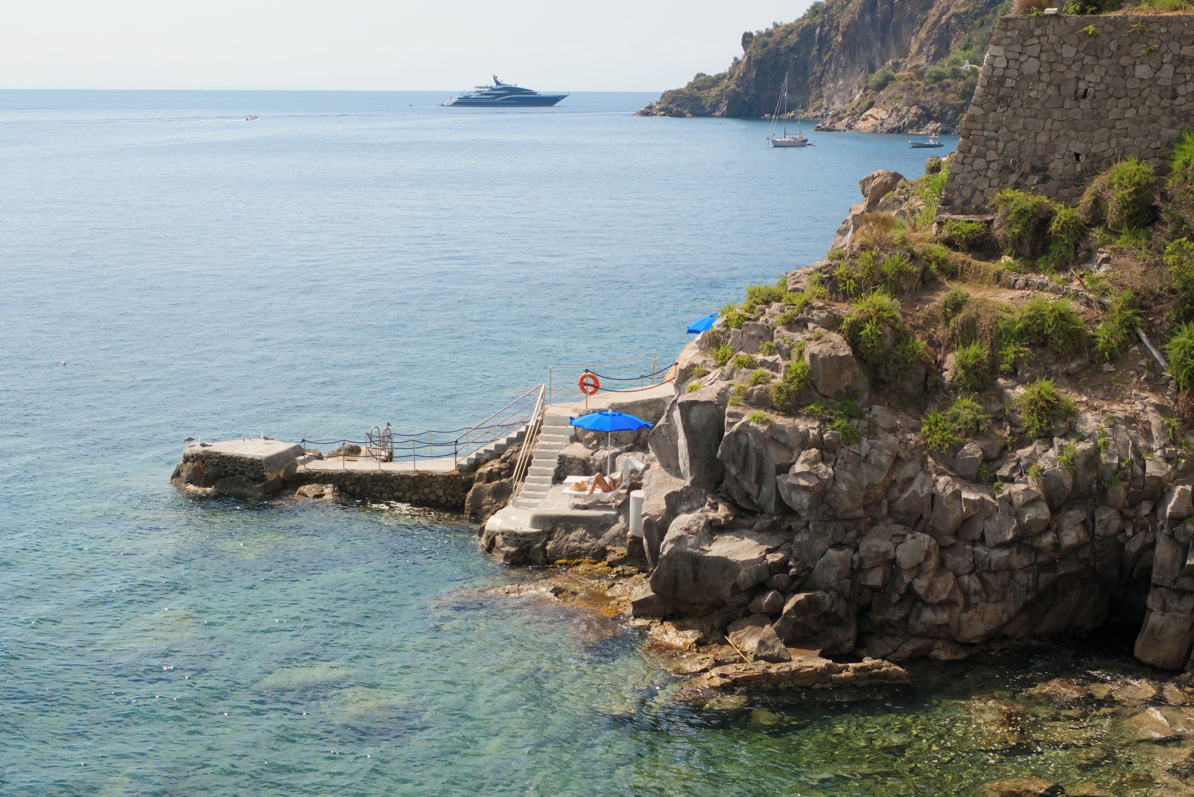 A coastal scene with a clear blue sea, rocky cliffs, and a small pier with steps leading into the water.
