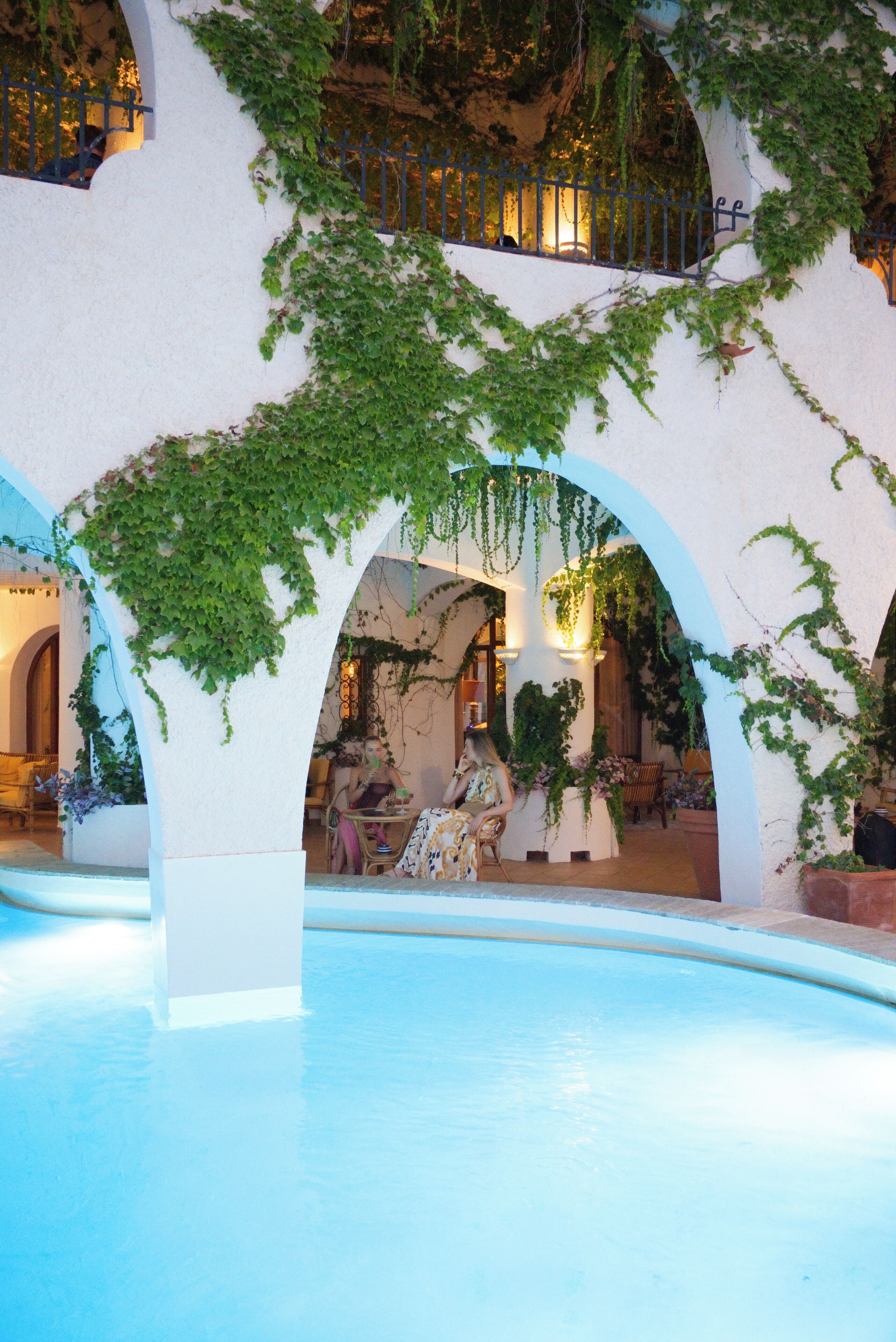 A pool area with a white wall and arches, surrounded by greenery.