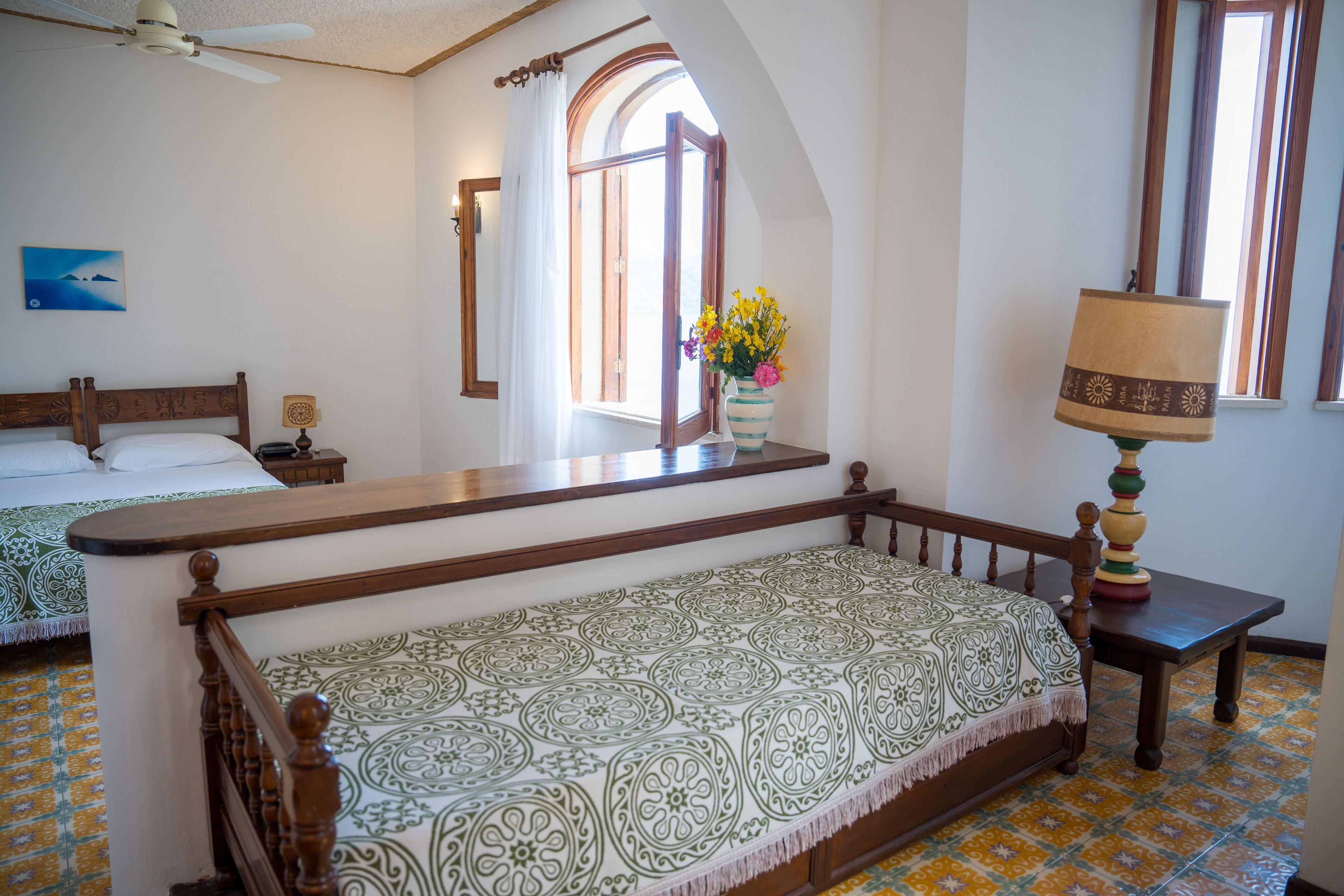 Un dormitorio con cama, un banco, una mesita de noche, una lámpara y una ventana con cortinas.