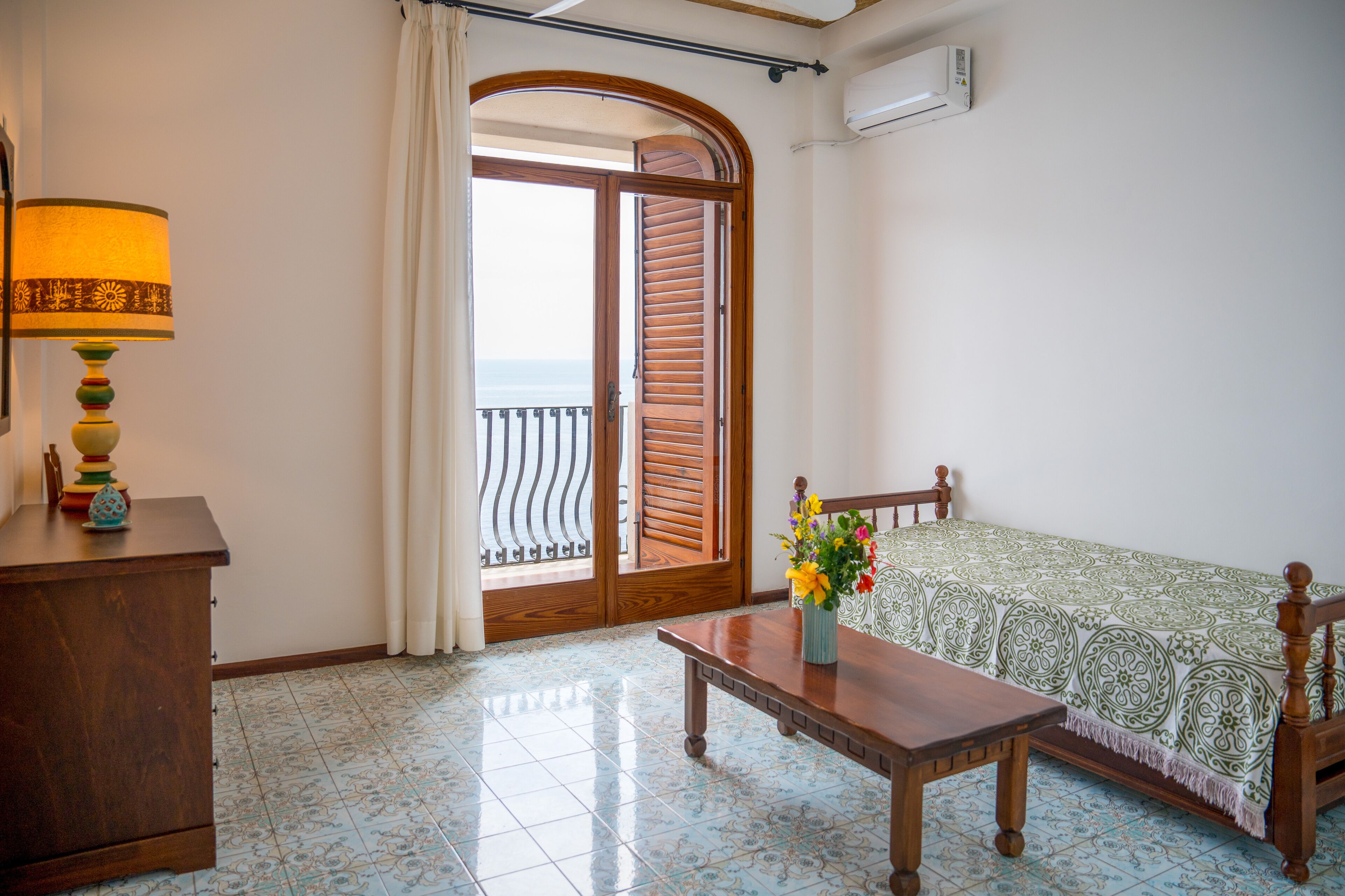 A room with a wooden bench, a table with a vase of flowers, a lamp, and a view of the sea through a door with wooden shutters.