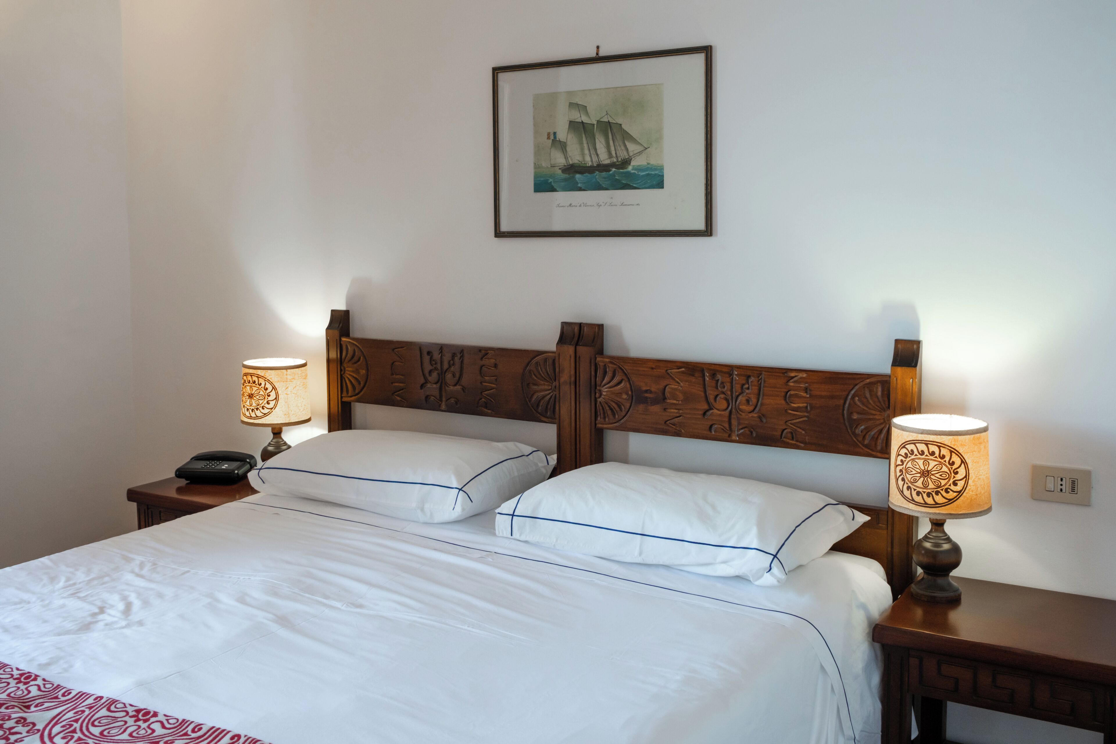 A hotel room with a wooden headboard, two bedside lamps, a framed picture on the wall, and a neatly made bed with white linens.