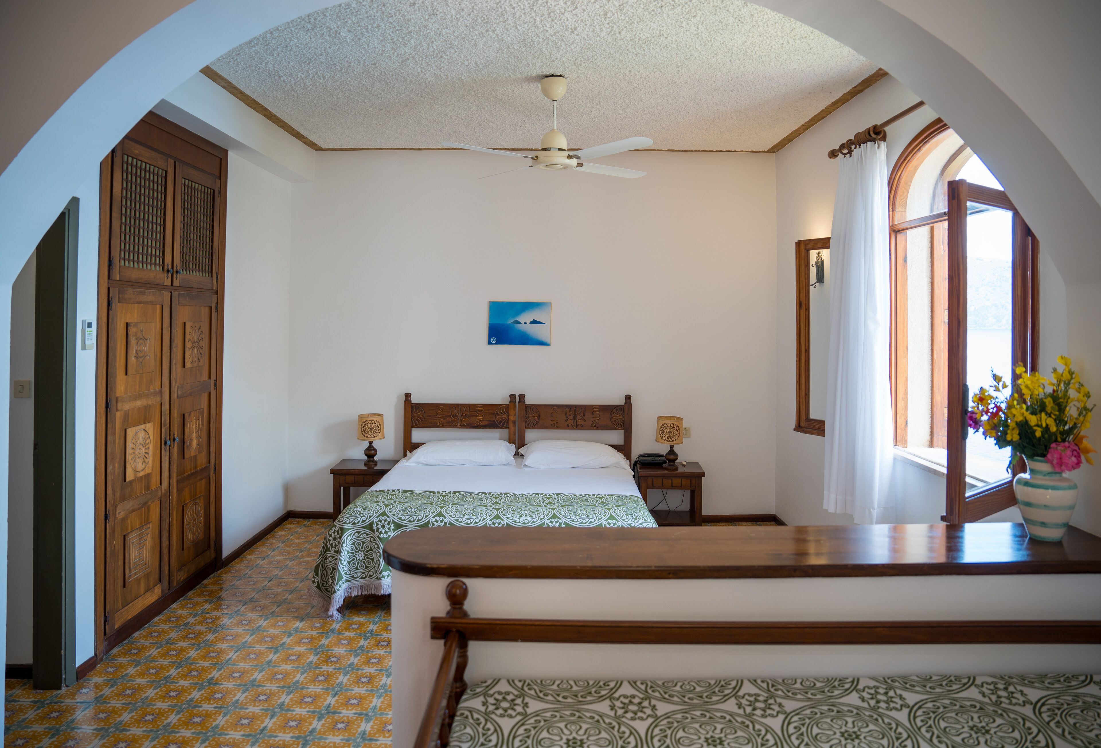 A bedroom with a bed, wooden wardrobe, a ceiling fan, a painting, a nightstand, a bedside table, a window with curtains, and a vase with flowers.