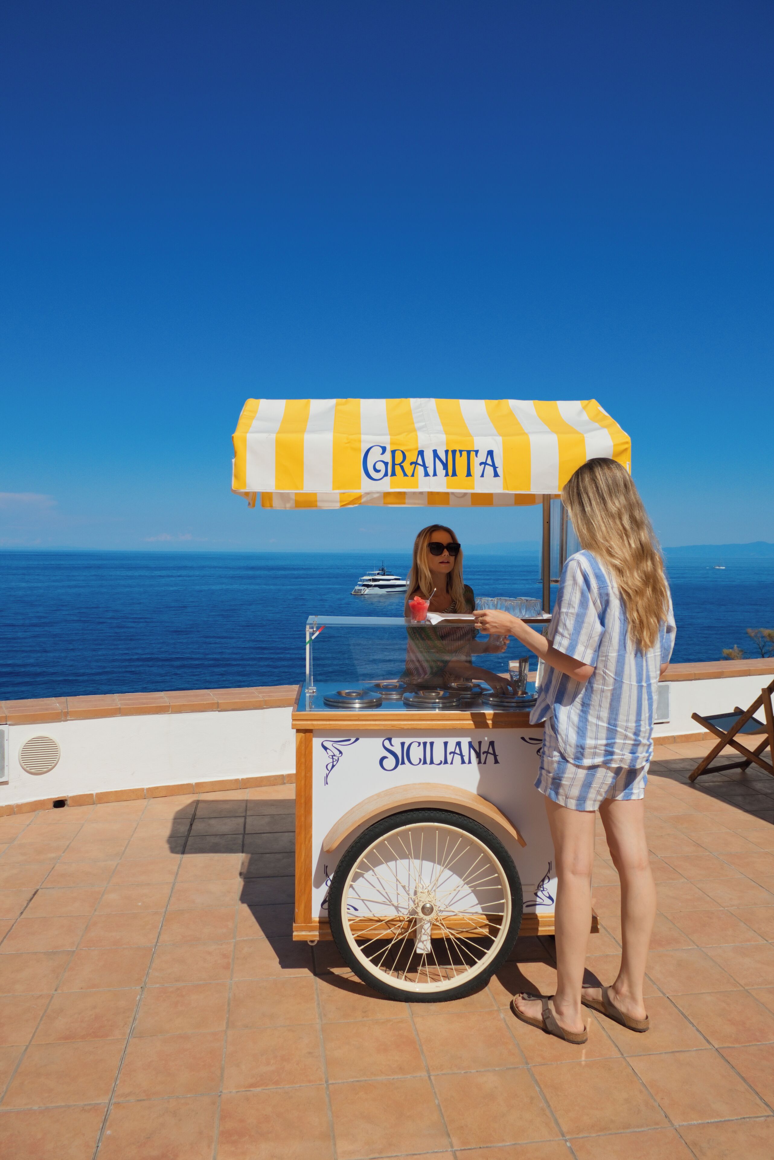 Un stand de restauration mobile, doté d’un auvent rayé sur lequel on peut lire les mots « GRANITA » et « SICILIANA », où l’on sert des plats près de la mer.