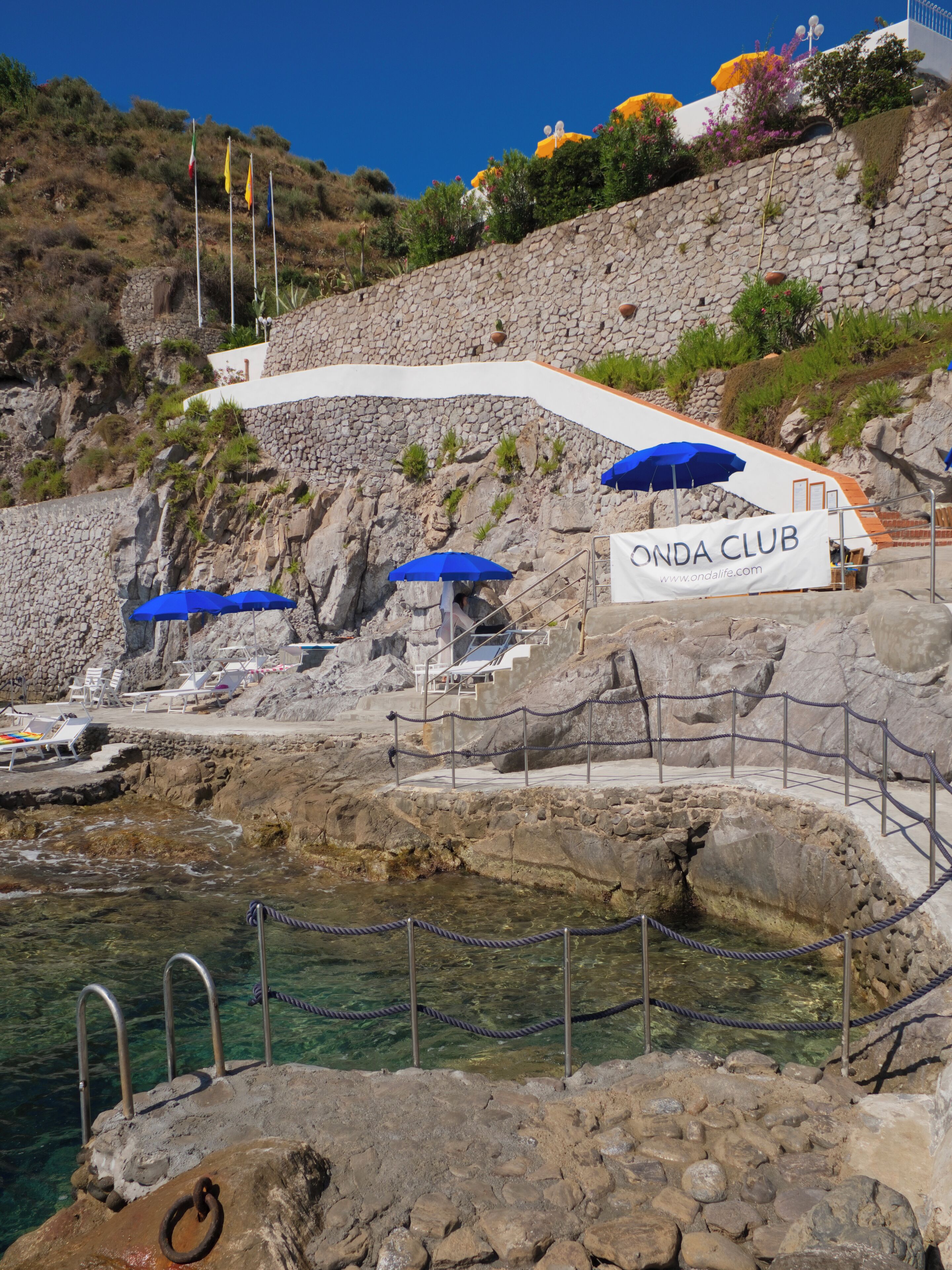 Eine felsige Küstenlandschaft mit einer Steintreppe, die zu einem weißen Gebäude mit der Aufschrift 'ONDA CLUB' führt. Blaue Sonnenschirme und freie Sicht auf das Meer.