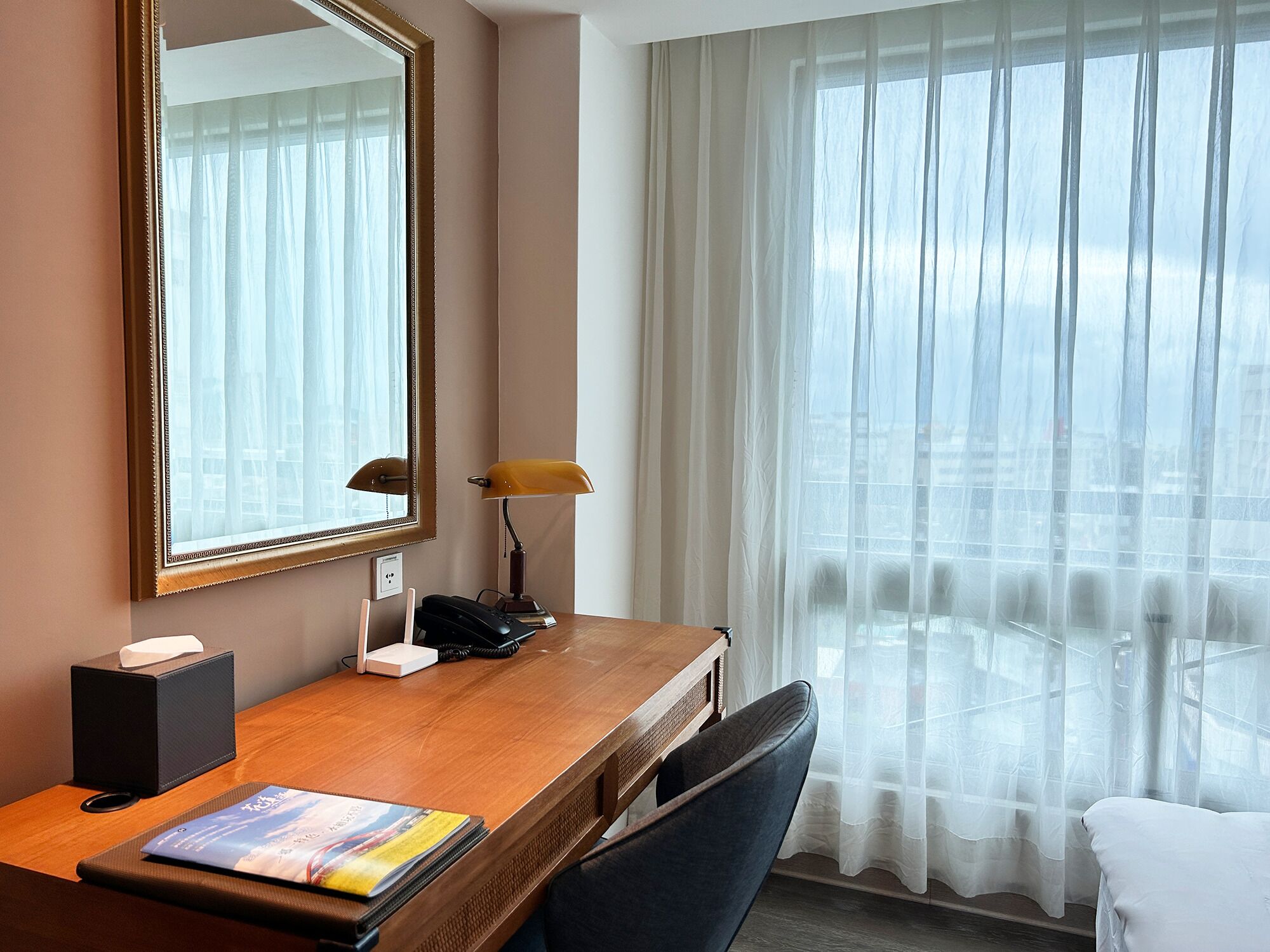 Habitación de hotel con un escritorio de madera, un teléfono, una lámpara, un espejo y una ventana con cortinas translúcidas.