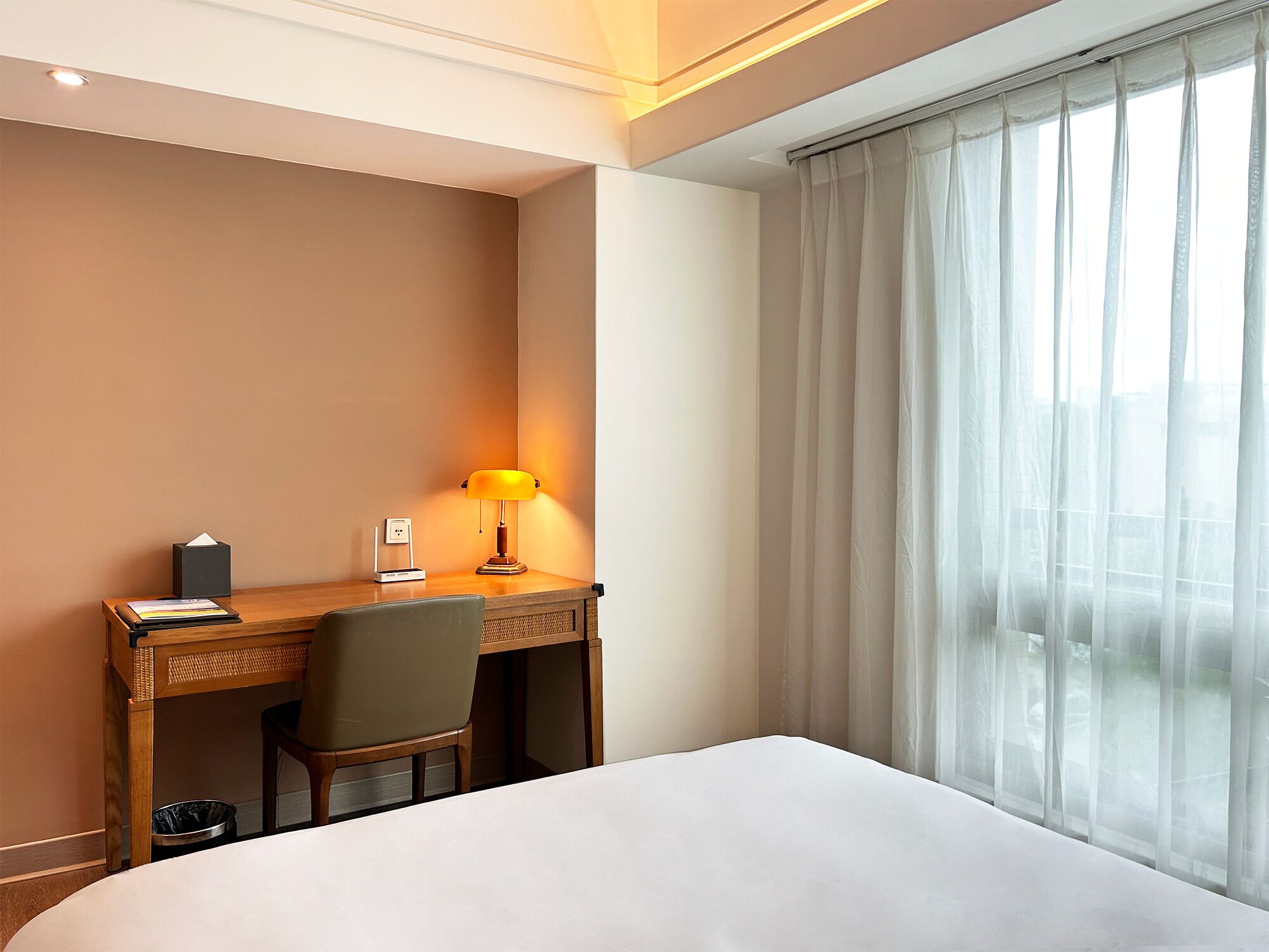 Habitación de hotel con cama, escritorio con lámpara, silla y ventana con cortinas translúcidas.