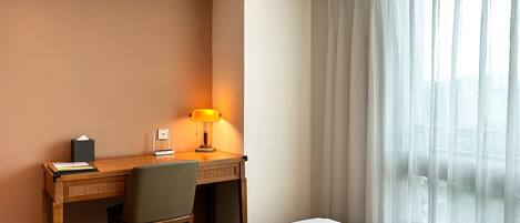 Habitación de hotel con cama, escritorio con lámpara, silla y ventana con cortinas translúcidas.