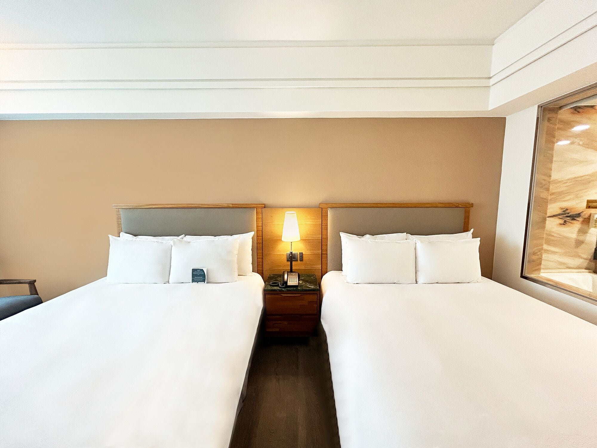A hotel room with two beds, a bedside table with a lamp, and a large window.