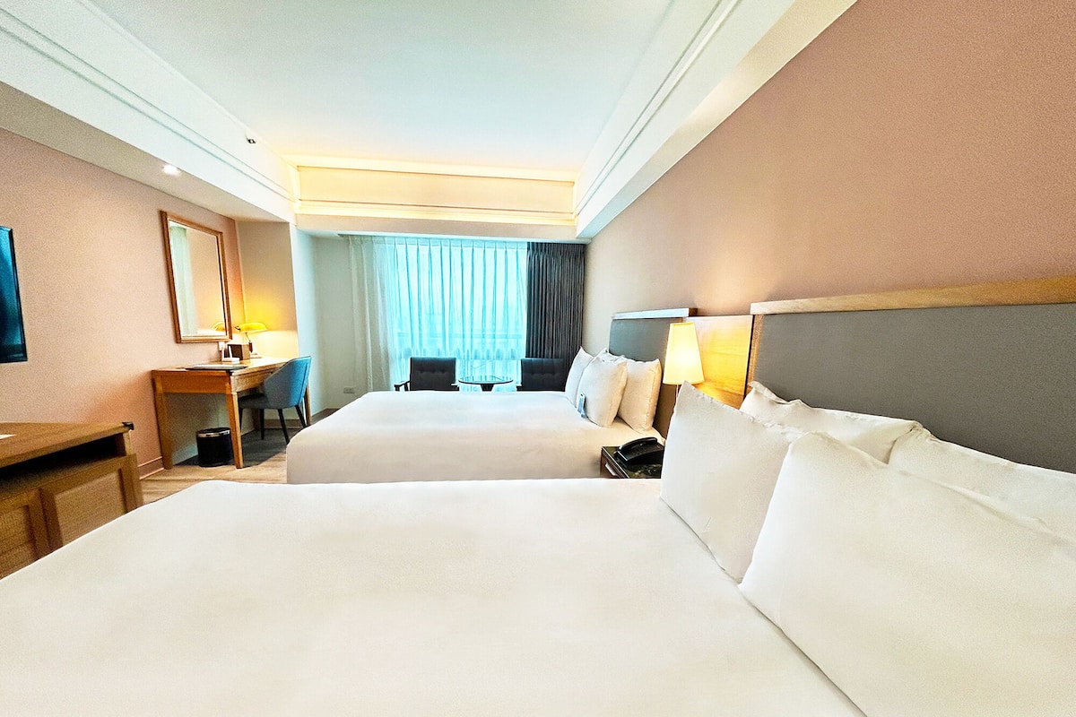 A hotel room with two beds, a desk, a chair, a lamp, and a large window with curtains.