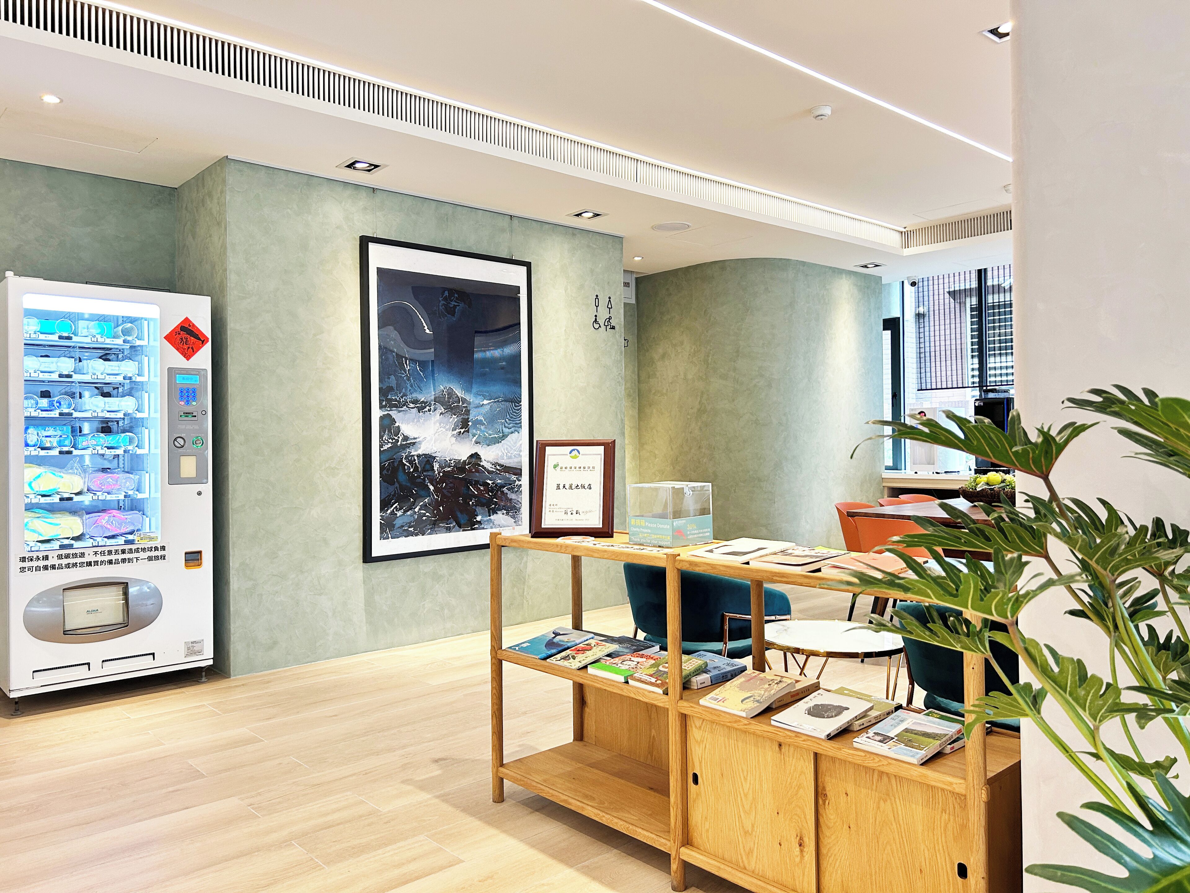 A modern interior with a vending machine, a framed artwork, a wooden desk with books, and a potted plant.