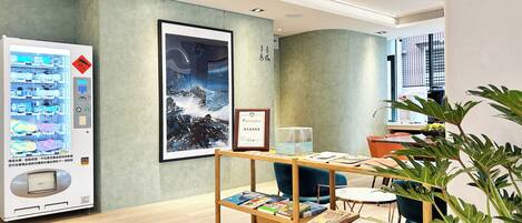 A modern interior with a vending machine, a framed artwork, a wooden desk with books, and a potted plant.