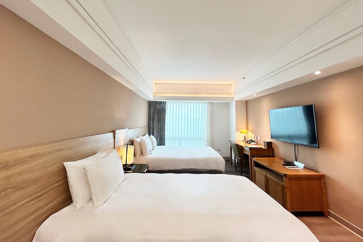 A hotel room with two beds, a desk, a television, and a window with curtains.