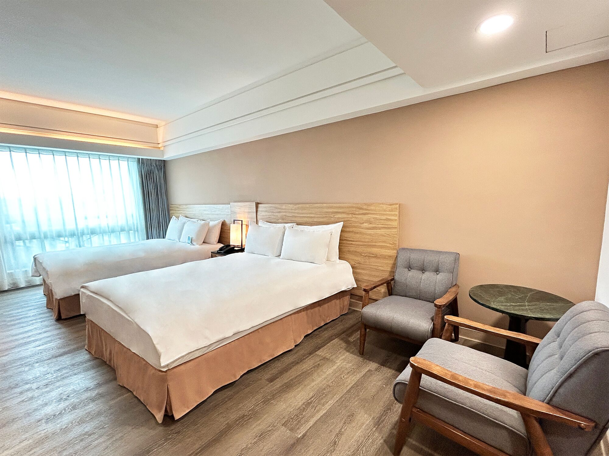 Ein Hotelzimmer mit zwei Betten, einem hölzernen Kopfteil, einem grauen Sessel, einem kleinen runden Tisch und einem großen Fenster mit Vorhängen.