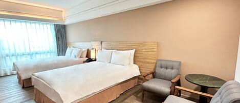 Ein Hotelzimmer mit zwei Betten, einem hölzernen Kopfteil, einem grauen Sessel, einem kleinen runden Tisch und einem großen Fenster mit Vorhängen.