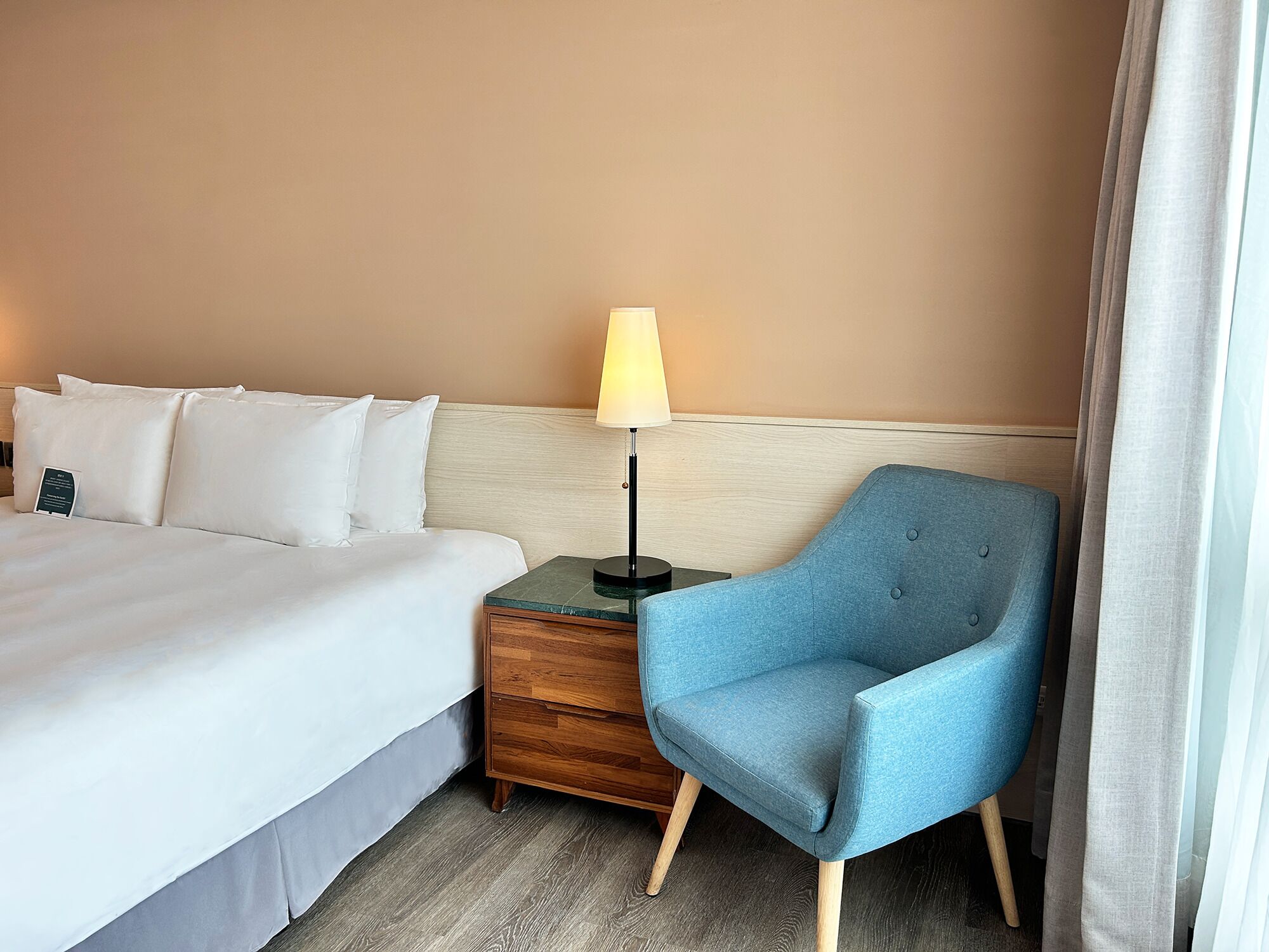 A hotel room with a bed, a nightstand, a lamp, a chair, and a window with curtains.