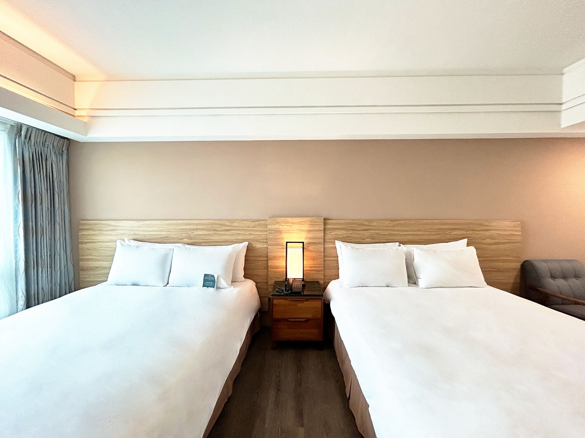 A hotel room with two beds, a nightstand, a lamp, and a chair.