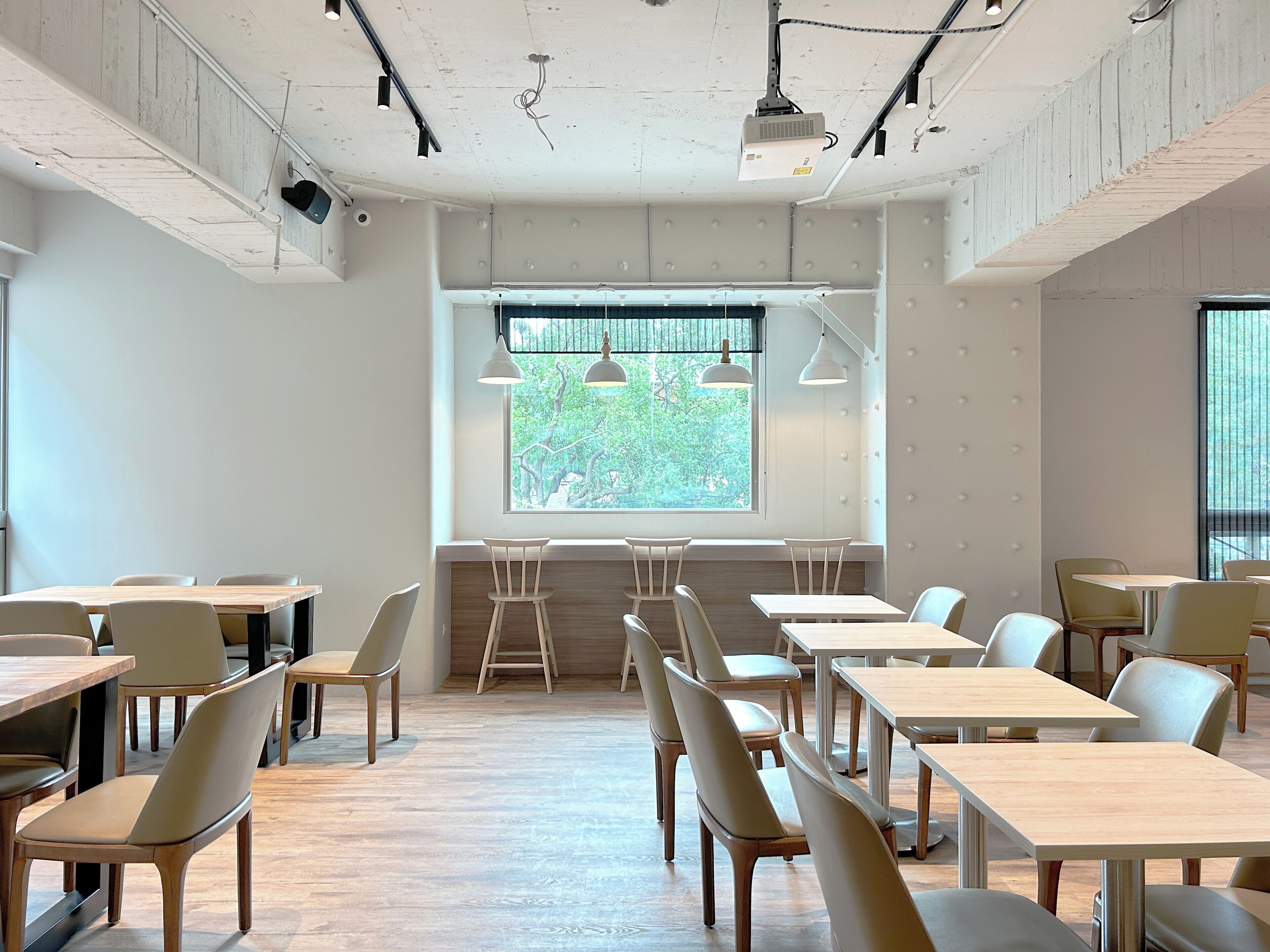 A modern dining area with wooden tables and chairs, a large window with a view of trees, and pendant lighting.