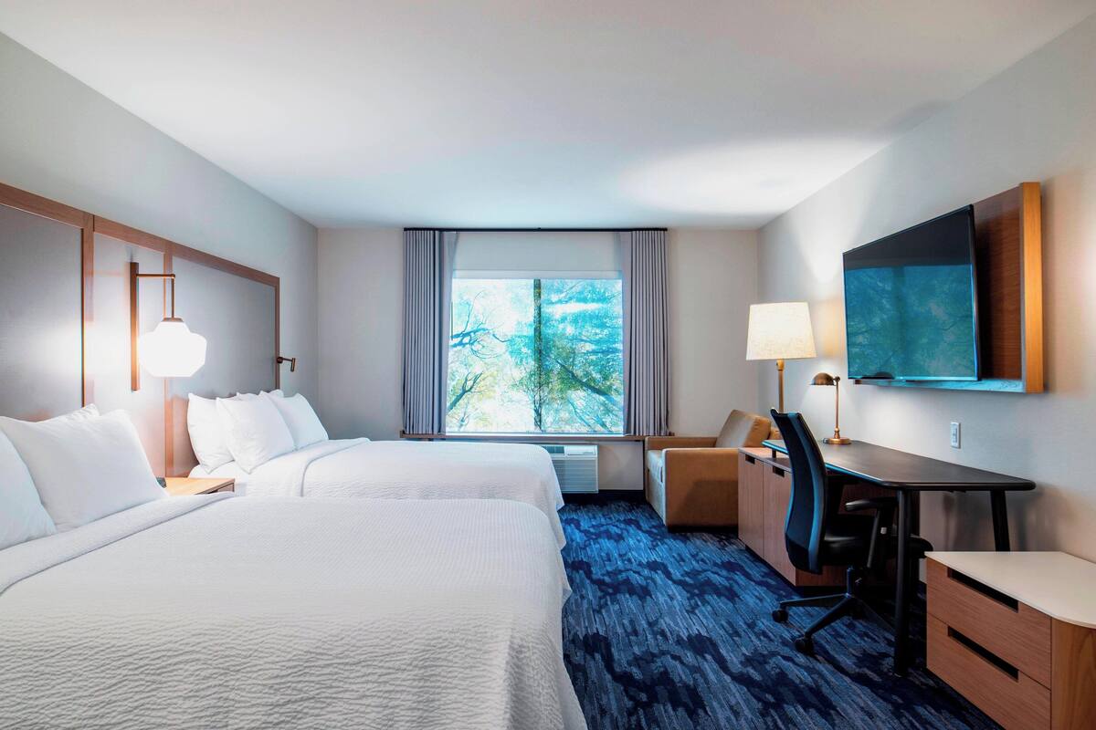 Une chambre d’hôtel avec deux lits, un bureau, une chaise, une télévision et une fenêtre donnant sur des arbres.