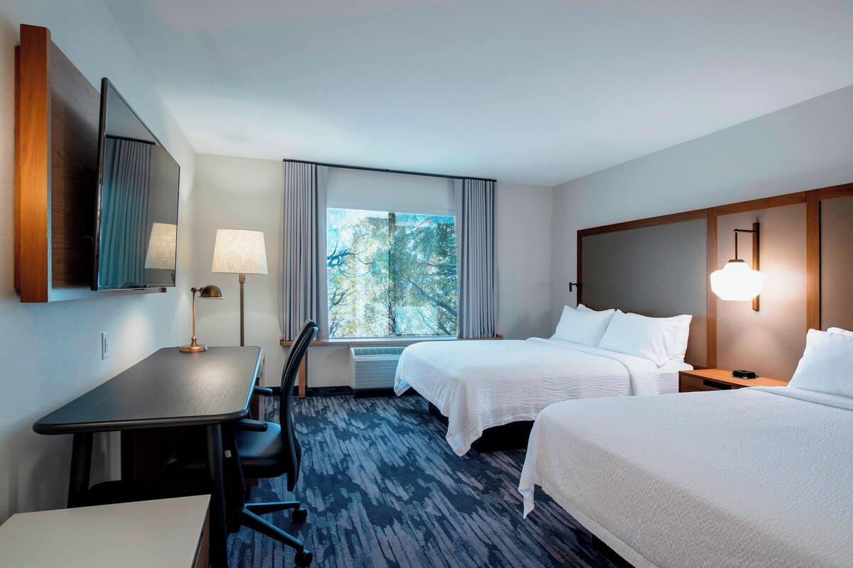 Une chambre d’hôtel avec deux lits, un bureau, une chaise, une lampe, une grande fenêtre avec des rideaux et une vue sur les arbres.