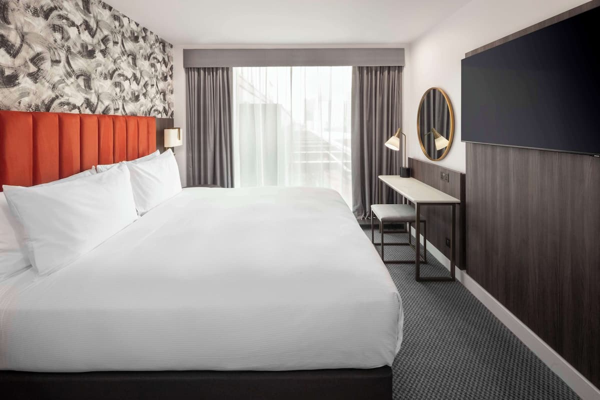 Ein Hotelzimmer mit einem großen Bett, einem Schreibtisch, einem Sessel, einem Fernseher und Blick auf eine Stadtlandschaft durch das Fenster.