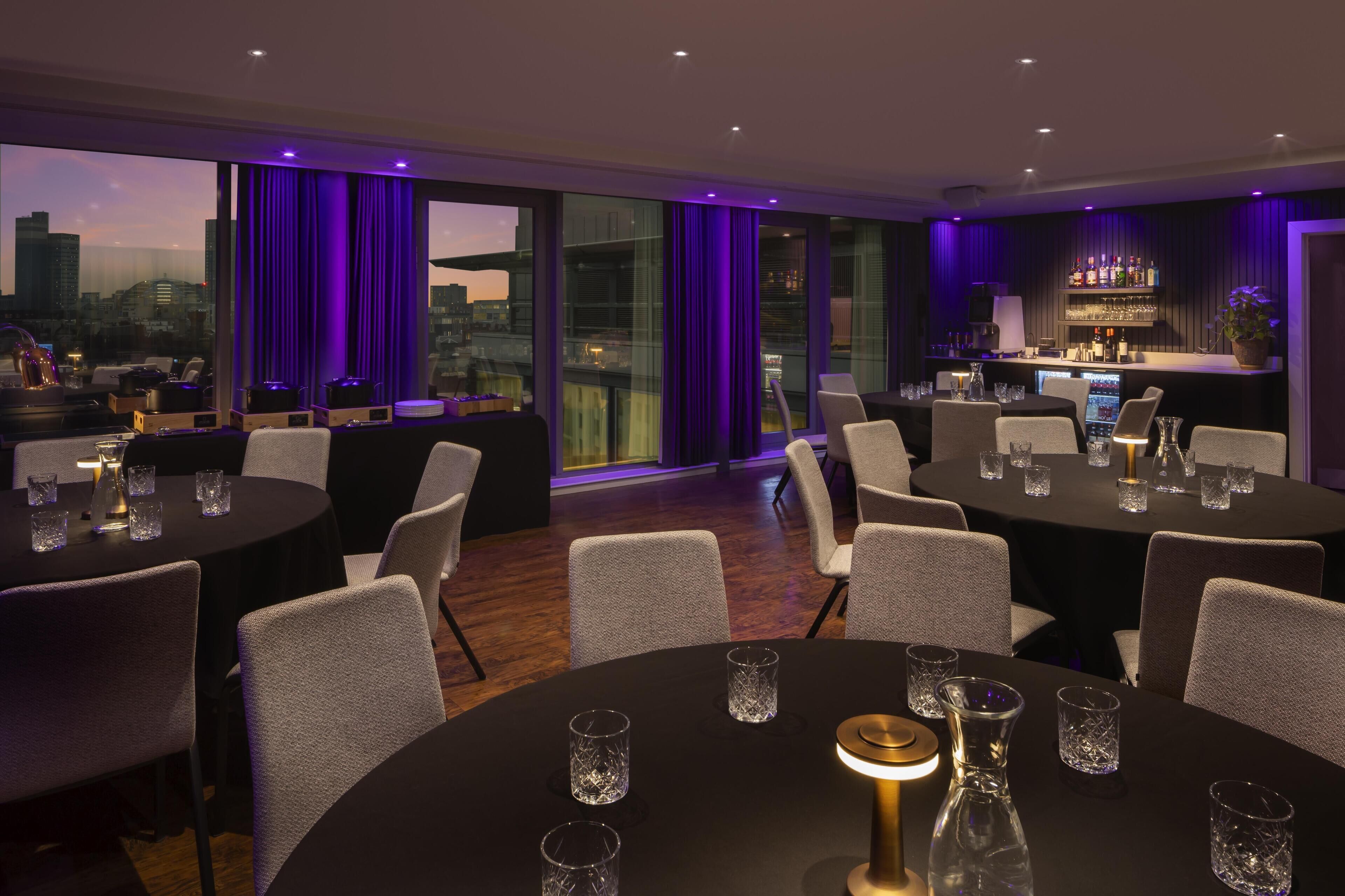 A modern dining area with round tables, chairs, and a bar with a city view.