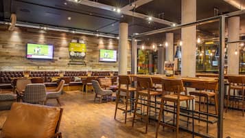 A modern bar area with wooden flooring, leather seating, and bar stools. There are two televisions mounted on the wall displaying sports.